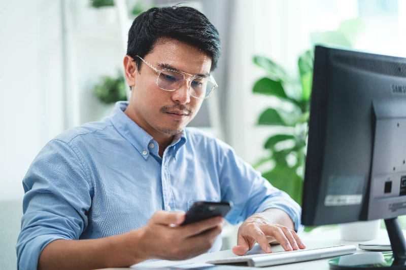 A man wearing glasses is sitting at a desk with a computer monitor and a cell phone